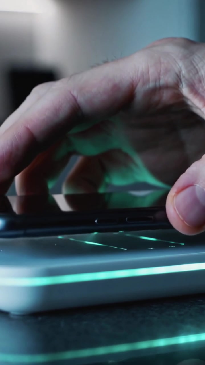 Vertical video: Entering hand placing phone onto green LED charging pad on counter, starting charge