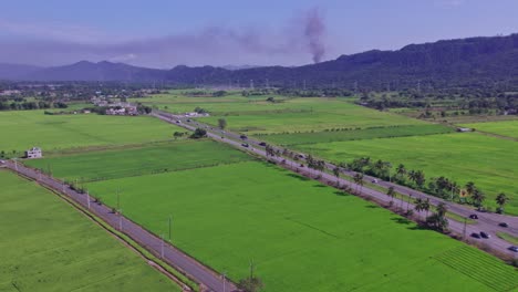 Vista-Aérea-De-La-Conducción-De-Camiones-En-La-Carretera-Al-Lado-De-Los-Campos-Agrícolas-Y-Pilas-De-Humo-Detrás-De-Las-Montañas-De-La-Fábrica-Industrial-En-El-Fondo---Concepto-De-Contaminación-Del-Aire