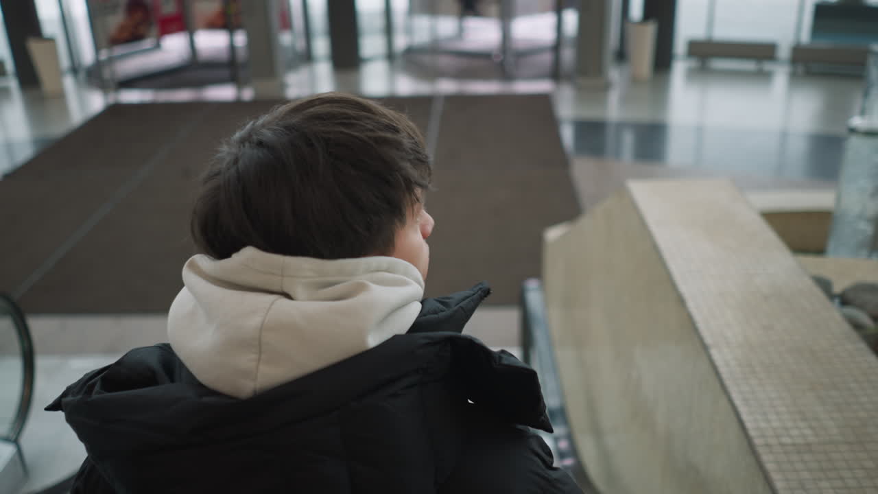 Teenager wearing black hoodie jacket descending escalator mall, focused expression, motion blur on steps and urban casual style, soft light, background shoppers blurred, travel and leisure atmosphere