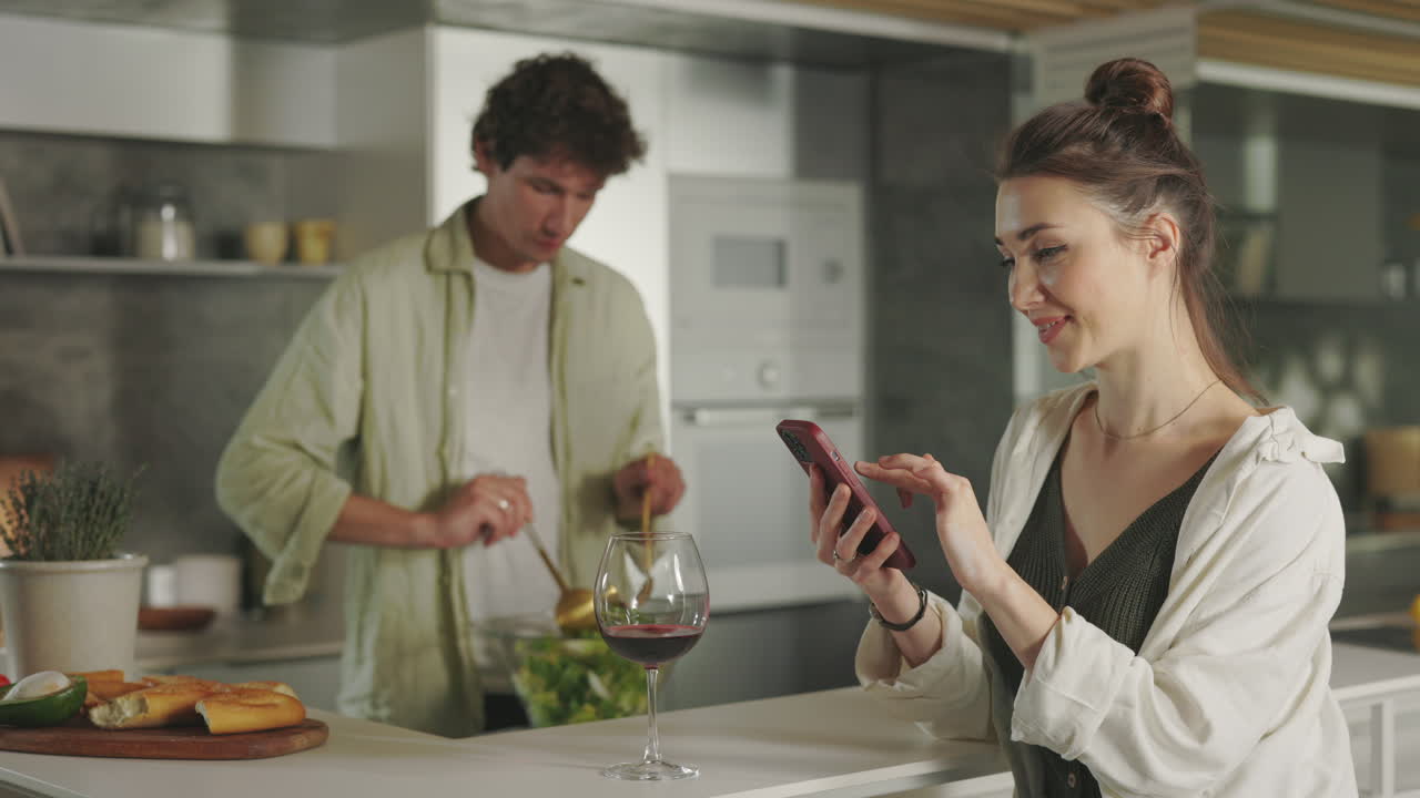 A couple in a modern kitchen, with one person stirring a salad and the other using a smartphone, a glass of red wine on the counter