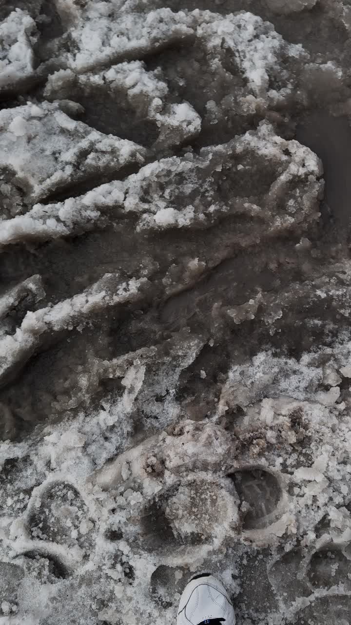 POV di un uomo che cammina con delle sneakers su un marciapiede urbano fangoso durante il disgelo di inizio primavera, alla ricerca di un modo per attraversare, ma bloccato da fango e pozzanghere sporche. Formato verticale