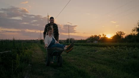 Ein-Glücklicher-Bauer-Schiebt-Seine-Freundin,-Einen-Bauern-In-Einem-Jeans-Overall,-In-Einer-Schubkarre-Am-Abend-Bei-Sonnenuntergang-Auf-Einem-Feld-Auf-Einem-Bauernhof