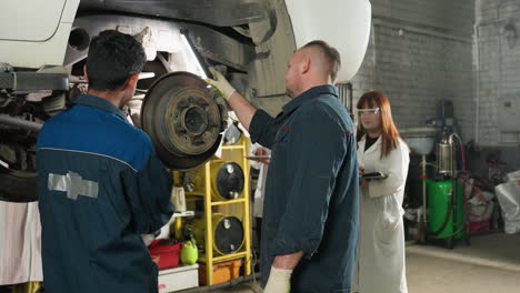 back view of man in blue uniform with lab scientists taking notes on tablet in mechanic workshop. industrial environment with tools, collaboration, research, and technical analysis in progress