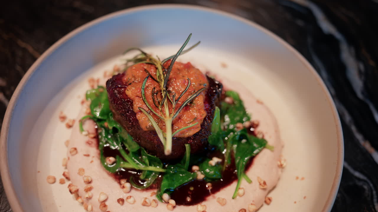 Close up of a gourmet meat dish served with sauce, greens, and rosemary garnish on a white plate