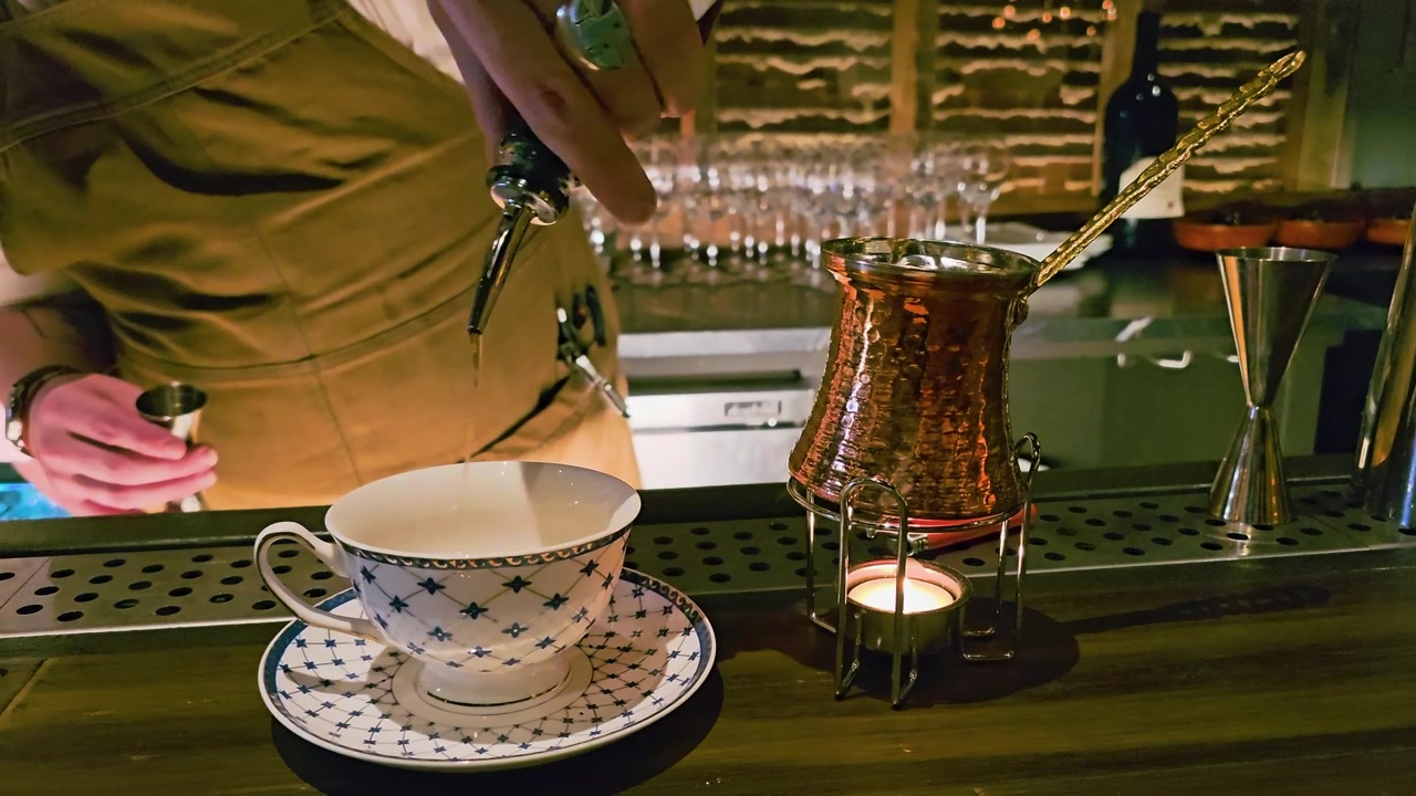 Stemningsfull barscene med en bartender som tilbereder en varm drikke i en porselenskopp, ved hjelp av en dampstav ved siden av en kobbervarmer, levende lys, målebeger, glass og varm loungebelysning for cocktail.
