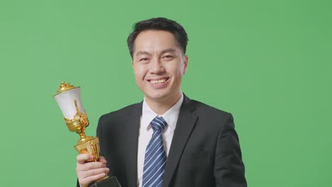 close up of asian business man in a suit and tie wears a gold medal and holds a gold trophy in his hand as the first winner smiling to camera on green screen background in the studio