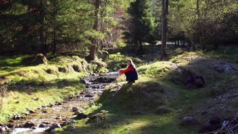 woman sitting next to flowing stream among meadows with bright sunlight in piatra craiului mountains, brasov county, romania, static shot