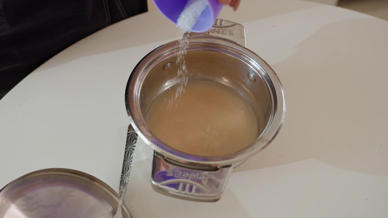 Primo piano dall'alto di una mano che versa un liquido trasparente da un misurino viola in un barattolo di vernice metallico aperto riempito con vernice beige, con un flusso visibile che crea increspature sulla superficie e un piccolo schizzo; mescolando con un oggetto metallico.