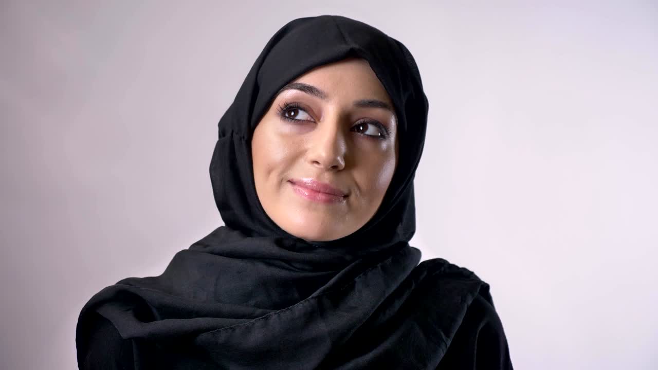 Young muslim girl in hijab opens eyes and watches at camera, smiling, religious concept, grey background