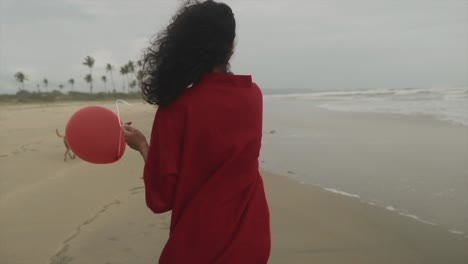 in this cinematic shot, a beautiful indian young girl wearing a vibrant red dress walks along the beach with a red balloon on her hand