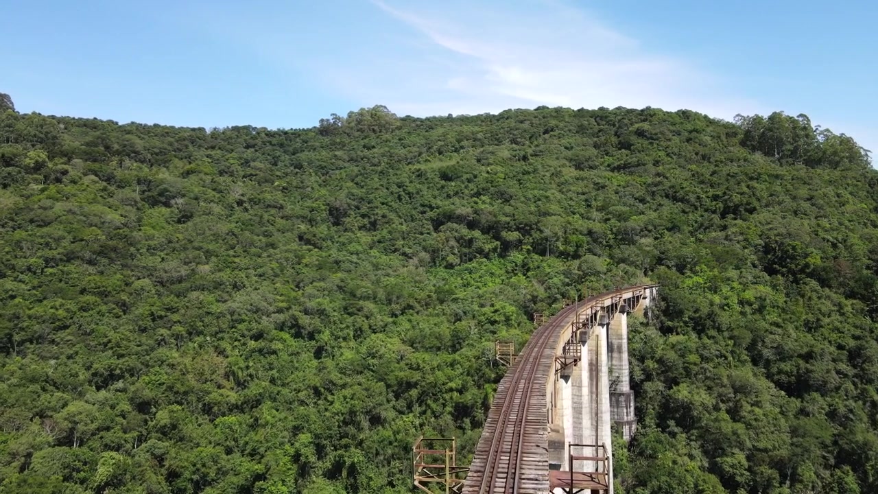 Traçado aéreo de uma ponte ferroviária alta atravessando um vale em direção a uma colina com floresta nativa.