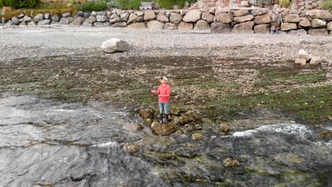 Asian-male-standing-on-a-rock-fly-fishing-in-the-Puget-Sound-wearing-a-red-shirt-and-rubber-boots-while-the-camera-pullsaway