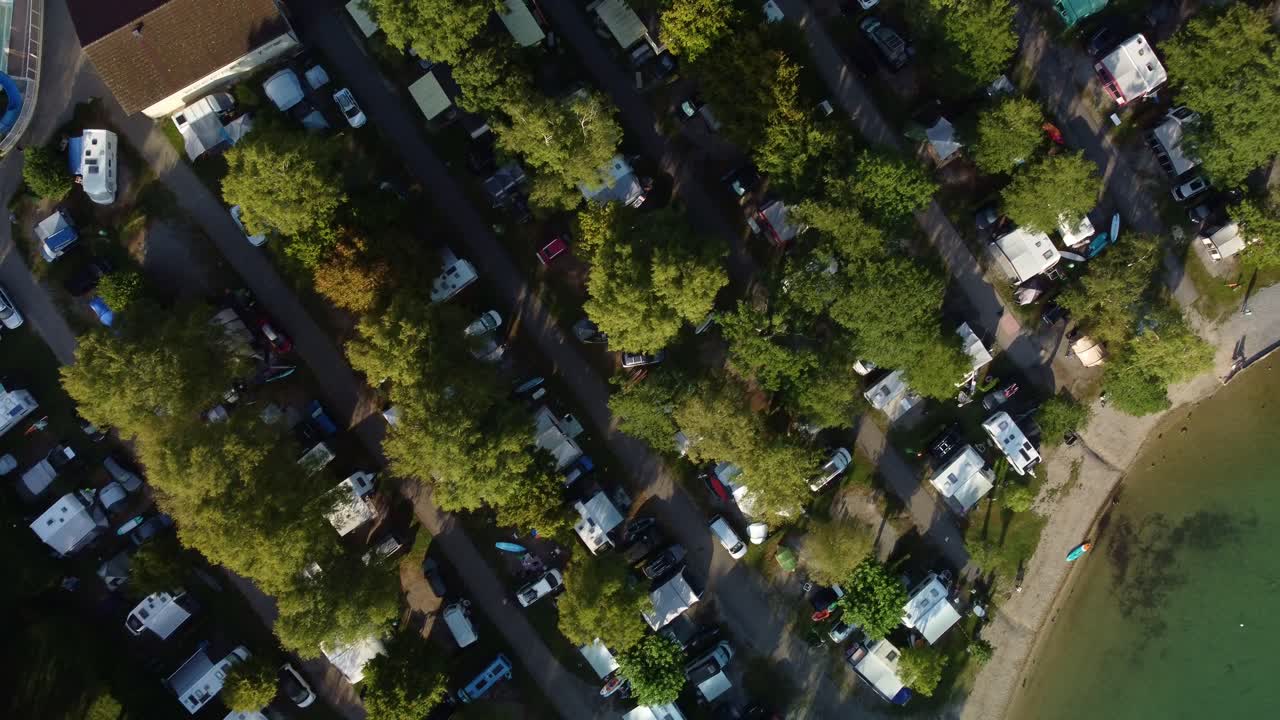 Top down aerial footage of Lac Bleu campsite and the bottom end of Lake Annecy in France. Footage moves to the right showing lines of pitches full of campers and a part of the beach and lake