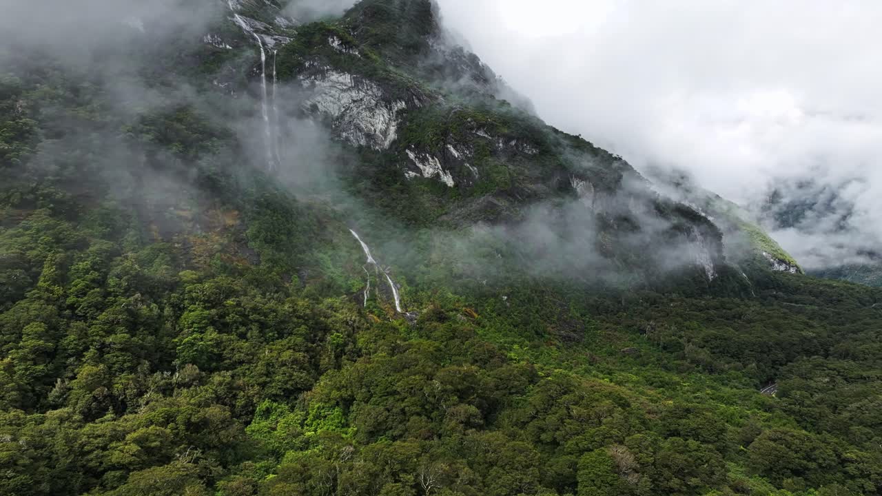 Światło słoneczne oświetla gęstą tropikalną dolinę z chmurami otulającymi wysoki wodospad w Milford Sound