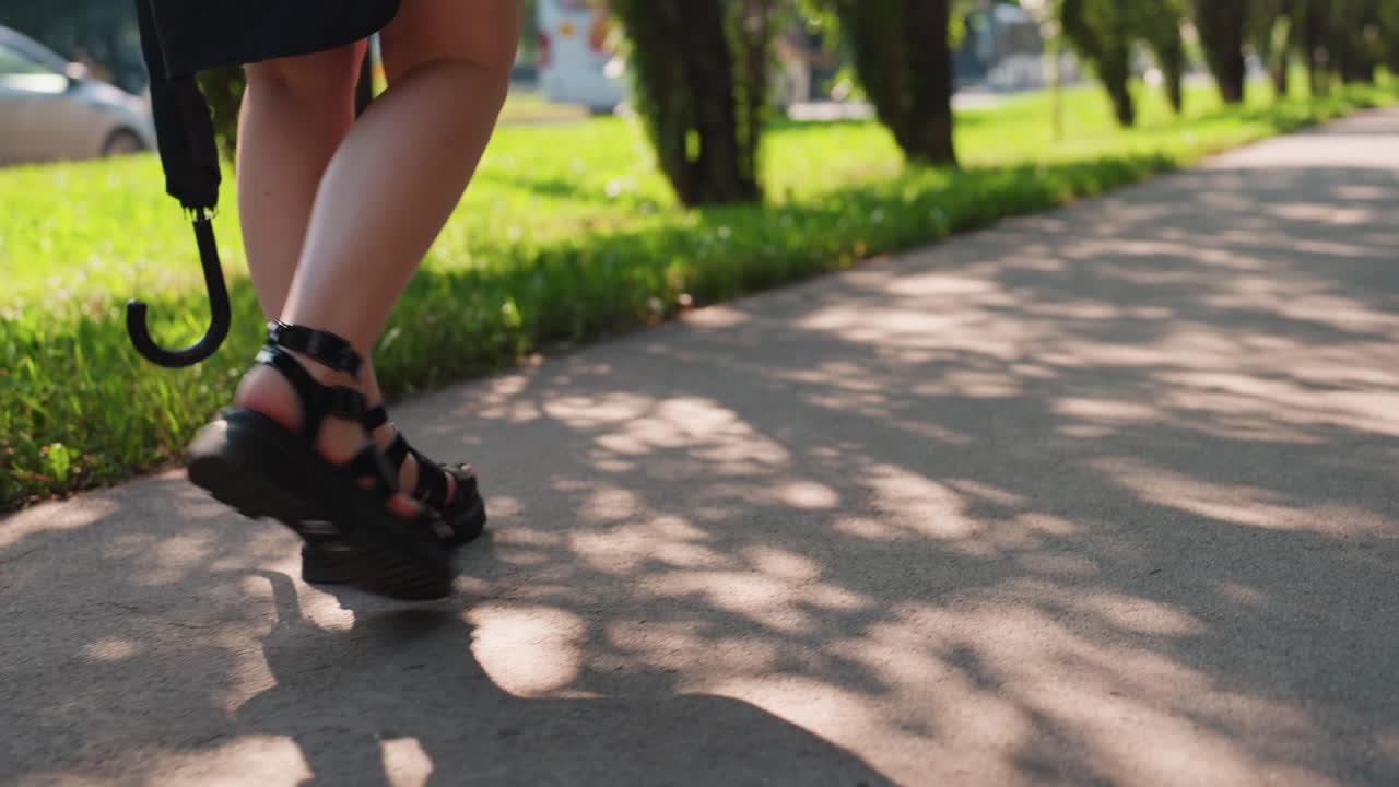 햇빛이 얼룩덜룩한 길을 따라 한 사람이 침착한 결심을 가지고 꾸준히 움직이며 걷고 있어요. 그늘진 숲길에서 편안한 샌들과 우산을 쓴 채 햇빛이 비치는 숲을 헤쳐나가는 모습이에요.