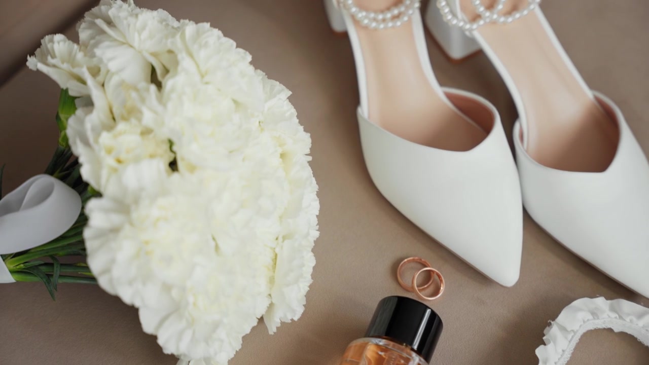 Chic close-up shot showcasing bridal white heels, pearl accents, wedding rings, floral bouquet, garter, and perfume bottle on a soft surface, perfect for wedding themes.