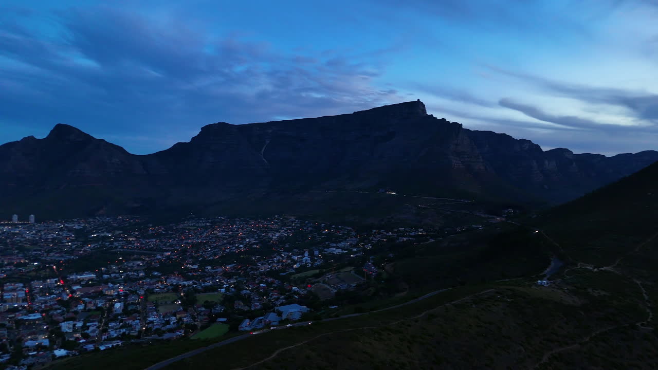 Cinematic aerial view of Table Mountain in Cape Town, South Africa, dramatic rocky cliffs with coastal cityscape at sunset, clouds rolling over peaks and golden evening light
