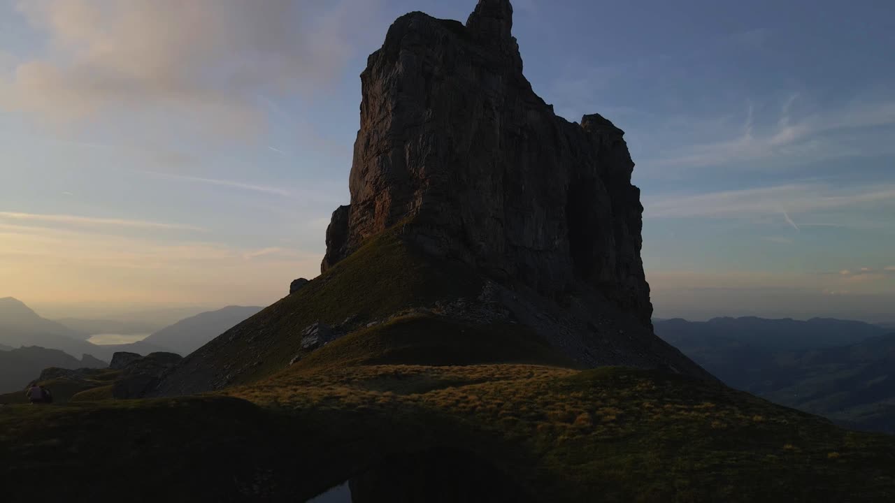Aerial view of the Gross Achslenstock rock formation at dawn, with a drone moving backward to reveal the rugged peak silhouetted against the soft morning sky, highlighting the serene alpine landscape
