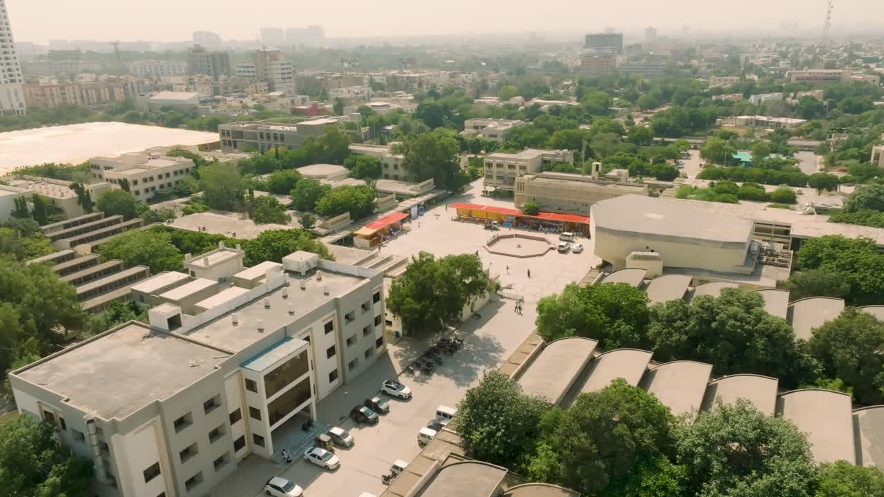 Drone pan over a central NED University campus building and adjacent parking lot filled with cars, with students gathered in the distance and hazy city skyline beyond