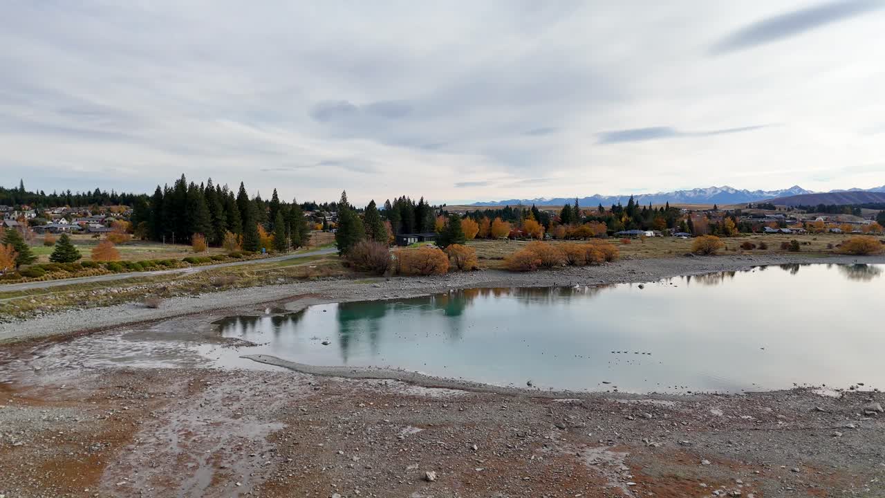 Drone footage captures Lake Tekapo's serene autumn landscape with reflective waters and vibrant foliage under overcast skies