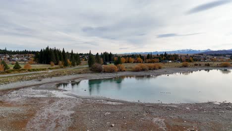 drone footage captures lake tekapo's serene autumn landscape with reflective waters and vibrant foliage under overcast skies
