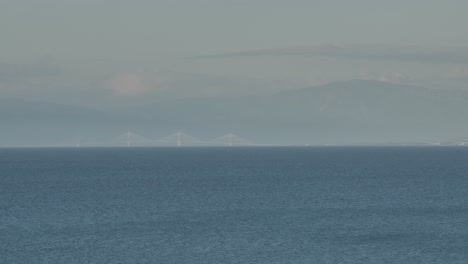 Langstrecken-Telebildschwenk-über-Eine-Weit-Entfernte-Brücke-Mit-Bergen-Im-Hintergrund-Und-Meer-Im-Vordergrund