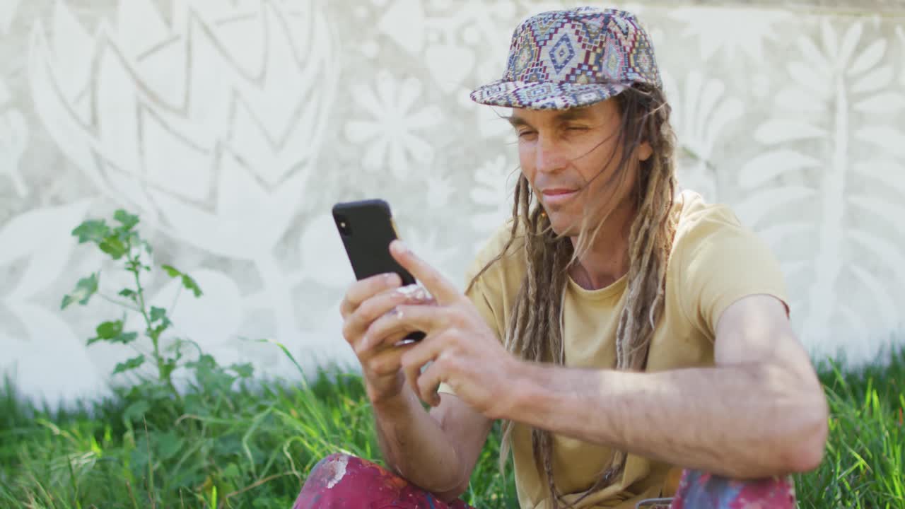 video de un hombre caucásico sonriente con rastas usando un teléfono inteligente sentado en la hierba por el mural en la pared