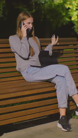 joven sentada en un banco al aire libre por la noche, multitarea con llamada telefónica y portátil, iluminada por la luz suave del parque, mochila roja a su lado en el banco, rodeada de vegetación