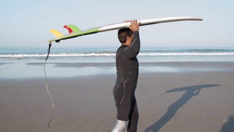 vista lateral de un surfista masculino con pierna protésica caminando en la playa con tabla de surf en la cabeza