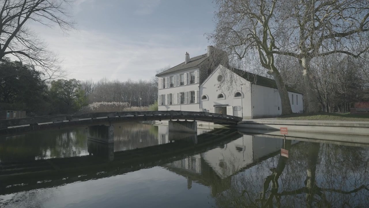 Una passerella di legno attraversa un laghetto calmo e riflettente accanto a un cottage bianco in un tranquillo parco parigino in inverno