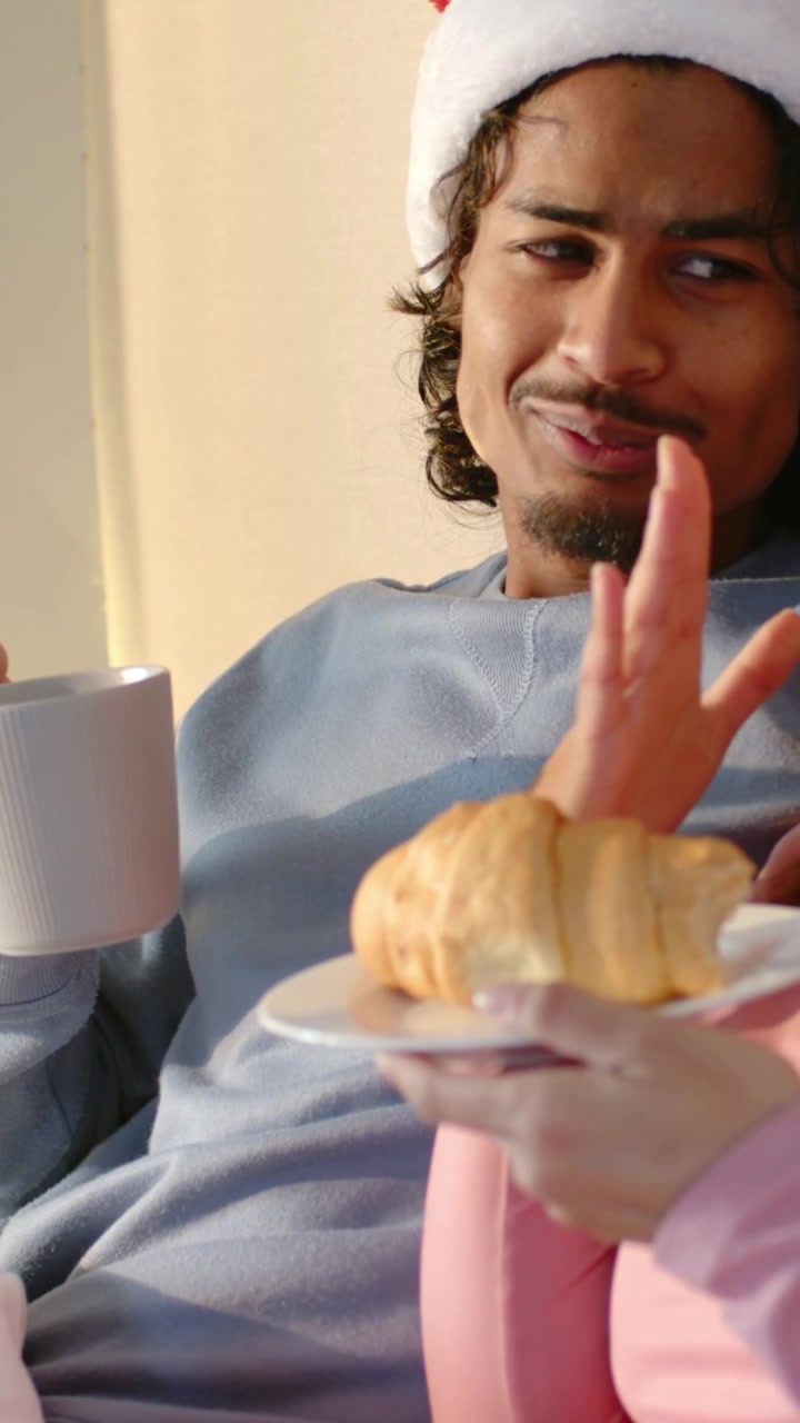 Video vertical: Hombre adulto ofrece un cruasán, lo prueba y luego lo rechaza en el sofá con una taza y un gorro de Papá Noel.