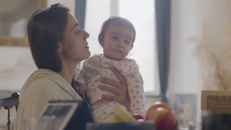 mujer feliz sosteniendo a su hija pequeña y hojeando una revista mientras se sienta en la mesa de la cocina por la mañana