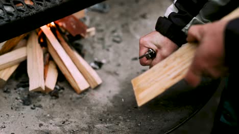 man cutting wood shavings of a log besides campfire