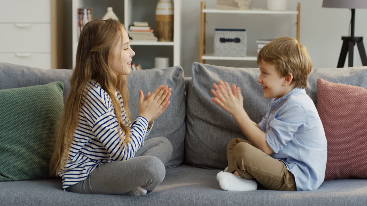 Two Small Cute Kids, Sister And Brother, Sitting On The Couch With Pillows In The Cozy Room And Playing A Game With Their Hands By Clapping Them