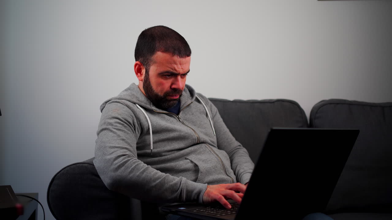 Bearded Man On Couch Thinking While Typing On Laptop Keyboard At Home