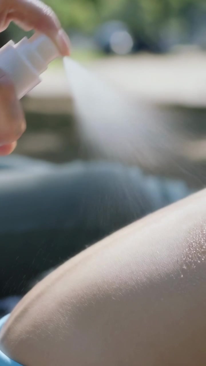 Vertical video: Pressing white pump, woman in blue top spraying exposed thigh at park for sun care