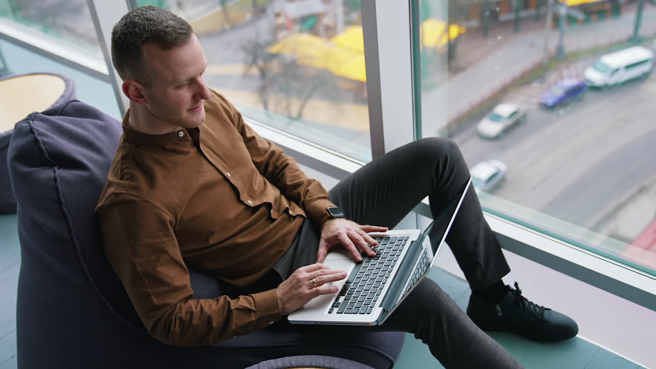 Handsome businessman in office by the window. Young man in brown shirt and trousers sitting on a comfortable ottoman and working on a laptop. City view background.