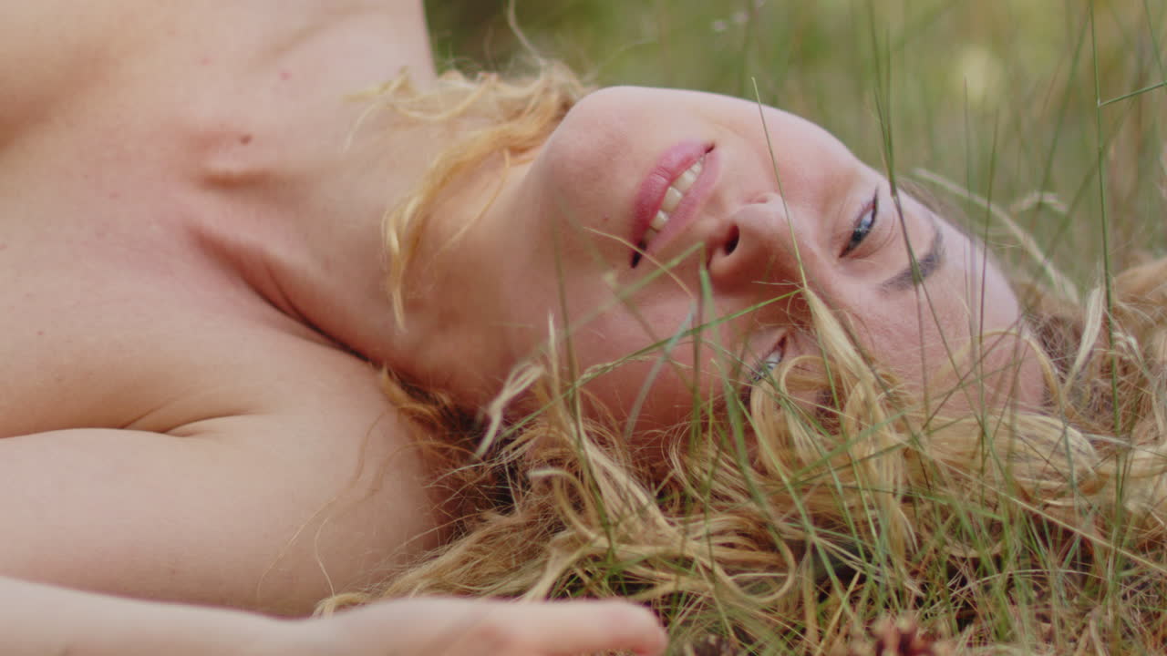 Close up of a beautiful woman peacefully lies in a sunny grass field and lovingly looks at the camera