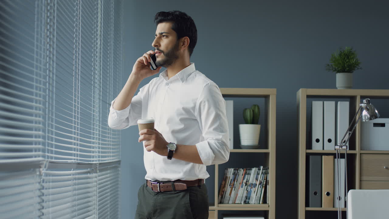 Young Handsome Stylish Arabian Businessman Standing At The Window In His Office, Talking Joyfully On The Phone And Drinking Coffee