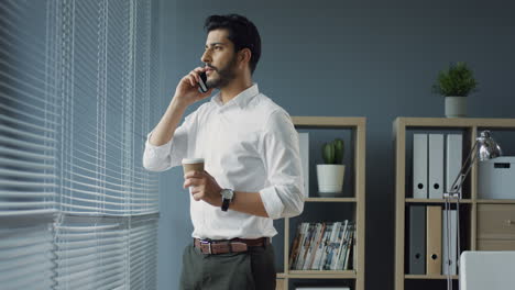 Young-Handsome-Stylish-Arabian-Businessman-Standing-At-The-Window-In-His-Office,-Talking-Joyfully-On-The-Phone-And-Drinking-Coffee