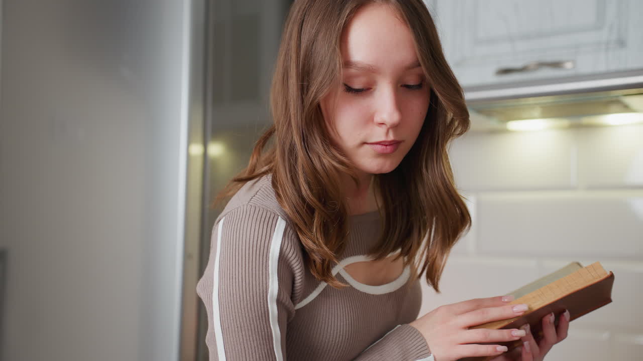 Una ragazzina legge un libro di cucina in cucina, si ferma per guardarsi intorno in modo contemplativo; sembra concentrata, forse sta pensando a una ricetta o alla preparazione di un pasto