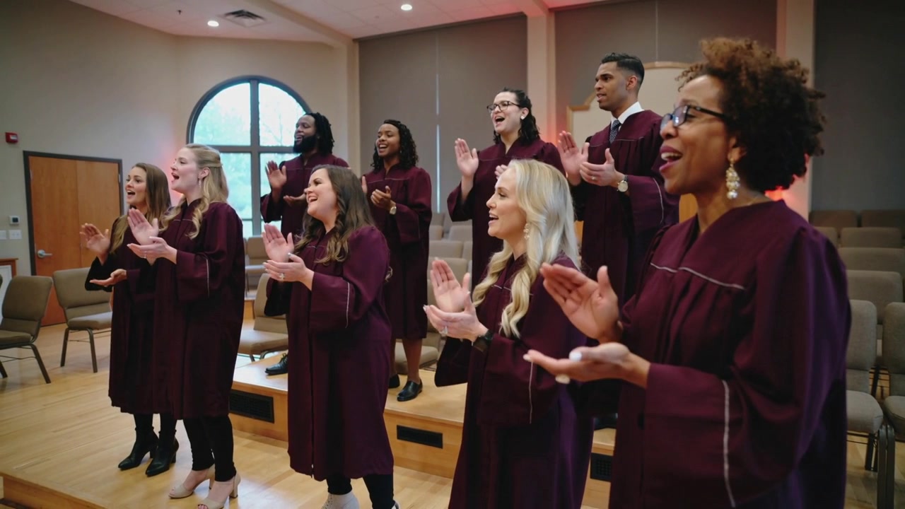 Al segnale musicale, un coro applaude e canta su una pedana della cappella, indossando tuniche color bordeaux.