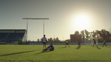 two soccer players sit on the field after practice as the sun sets behind the empty stadium lights