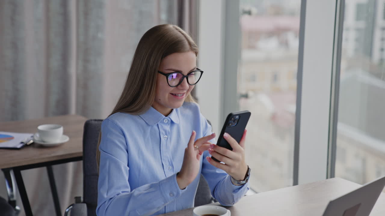 Young woman in office room sits at the desk. Lady in glasses is in front of laptop and typing message on her phone. Close up.