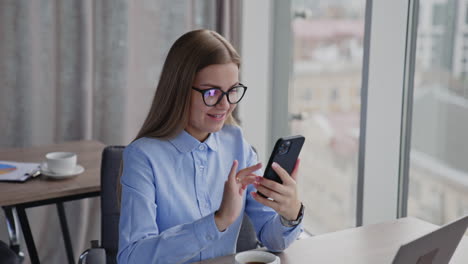 young woman in office room sits at the desk. lady in glasses is in front of laptop and typing message on her phone. close up.
