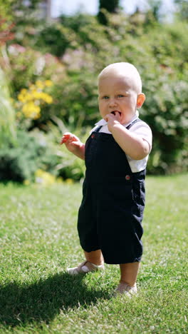 adorable caucasian blond baby boy in white t-shirt and black romper stands unsteadily on the grass. kid waves his hands and dances a little. vertical video.