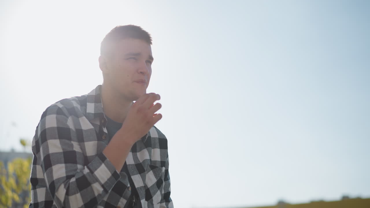 young man outdoors during sunny day puts sweet in mouth while sunlight creates soft glowing effect around him as he gently smiles with blurred buildings and trees in distant background
