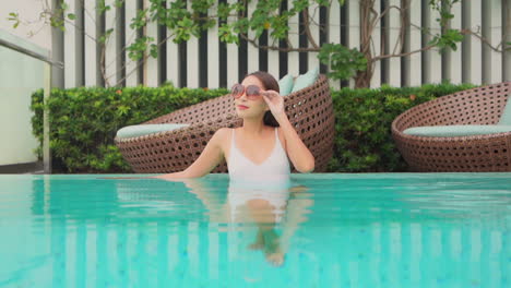 una mujer joven y bonita se ajusta las gafas de sol mientras se inclina contra el borde de la piscina