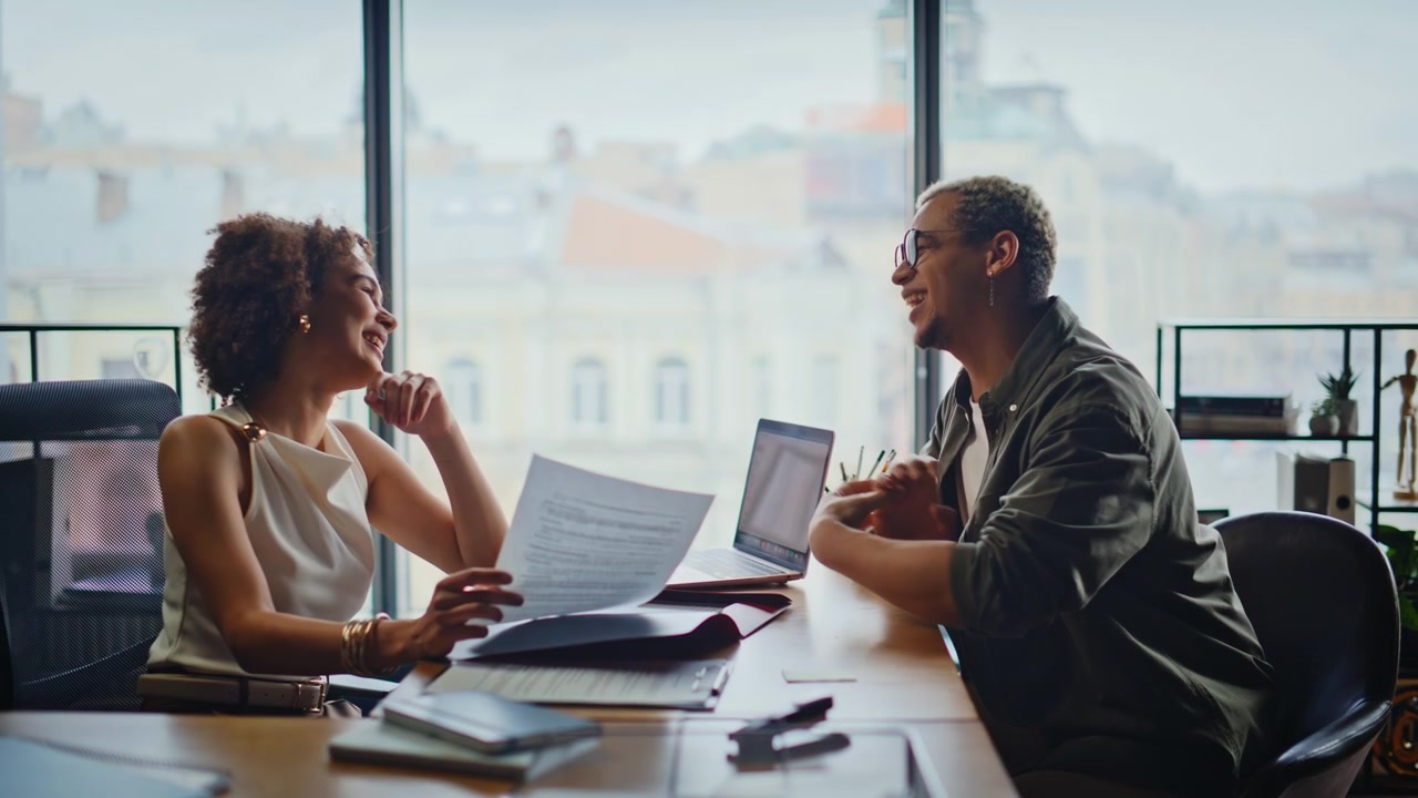 Two colleagues laughing office meeting. Woman reviewing documents coworker desk.