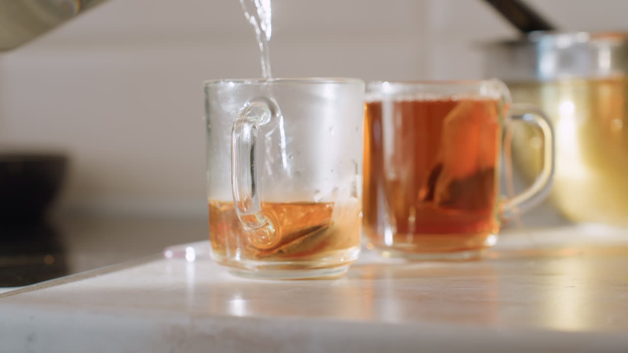 Close up pouring hot water into glass cup with teabag as liquid turns amber while tea steeps, with another cup already brewed in bright kitchen setting beside stainless steel bowl